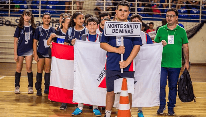 Delegação de Monte Santo de Minas entra na quadra com estudantes, bandeira da cidade e placa de identificação durante evento esportivo.