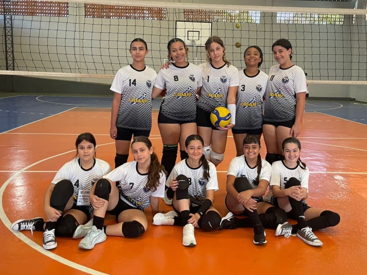 Time feminino juvenil de vôlei posa na quadra após partida, com algumas atletas em pé e outras sentadas.