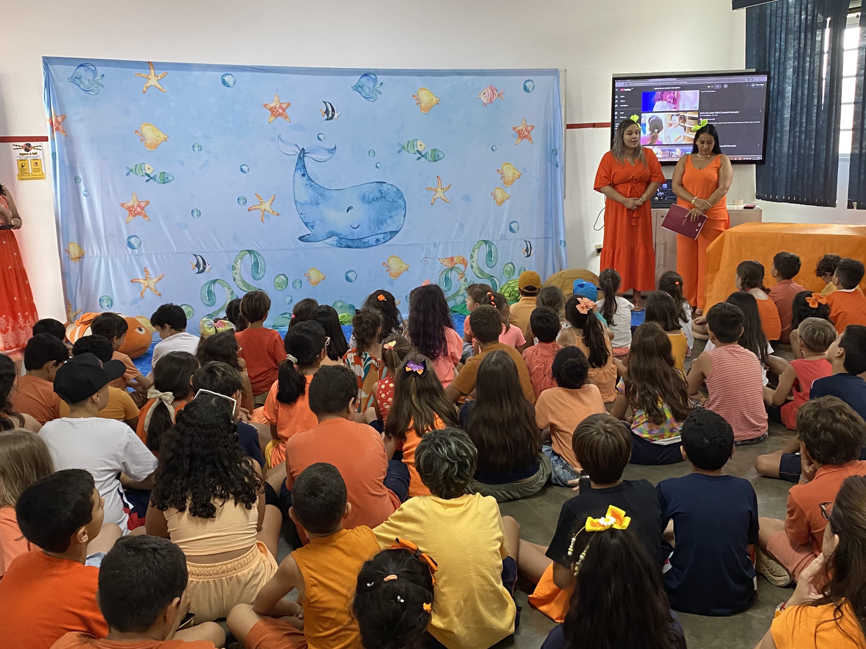 Crianças sentadas assistem a uma apresentação escolar com temática do fundo do mar, conduzida por duas educadoras.