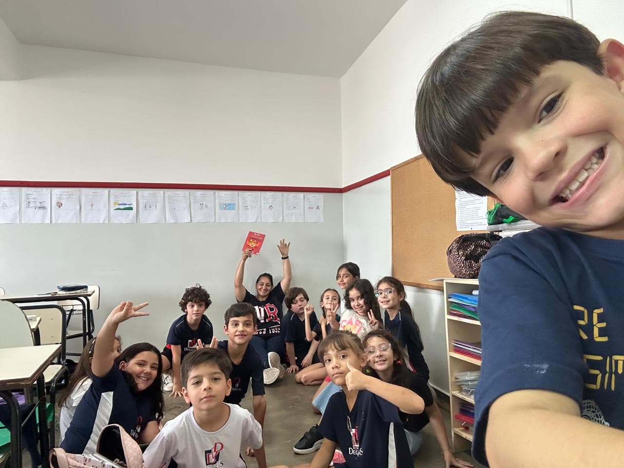 Turma de crianças reunidas em sala de aula, sorrindo e fazendo gestos para uma foto em grupo.