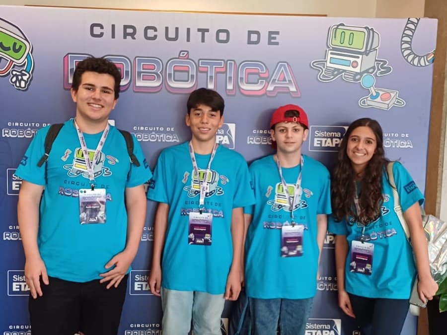 Quatro estudantes com camisetas e credenciais do Circuito de Robótica posando diante do painel do evento.