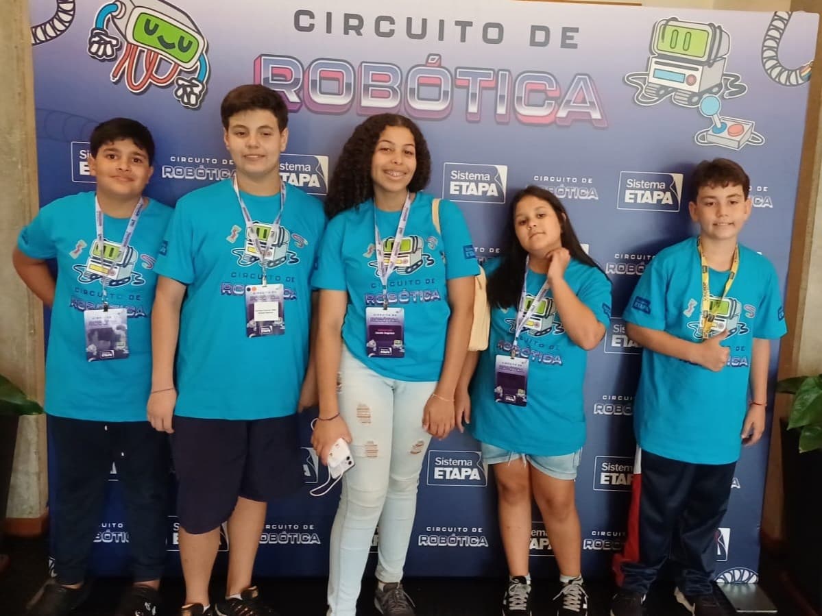 Cinco estudantes participantes do Circuito de Robótica posando em frente ao painel do evento do Sistema Etapa.