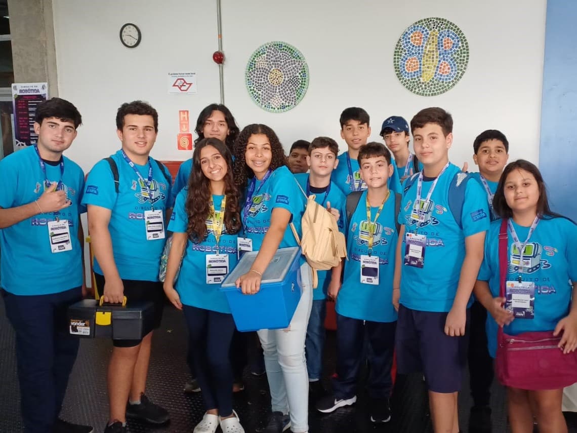 Grupo de estudantes com camisetas do Circuito de Robótica reunido durante o evento, carregando materiais e equipamentos.