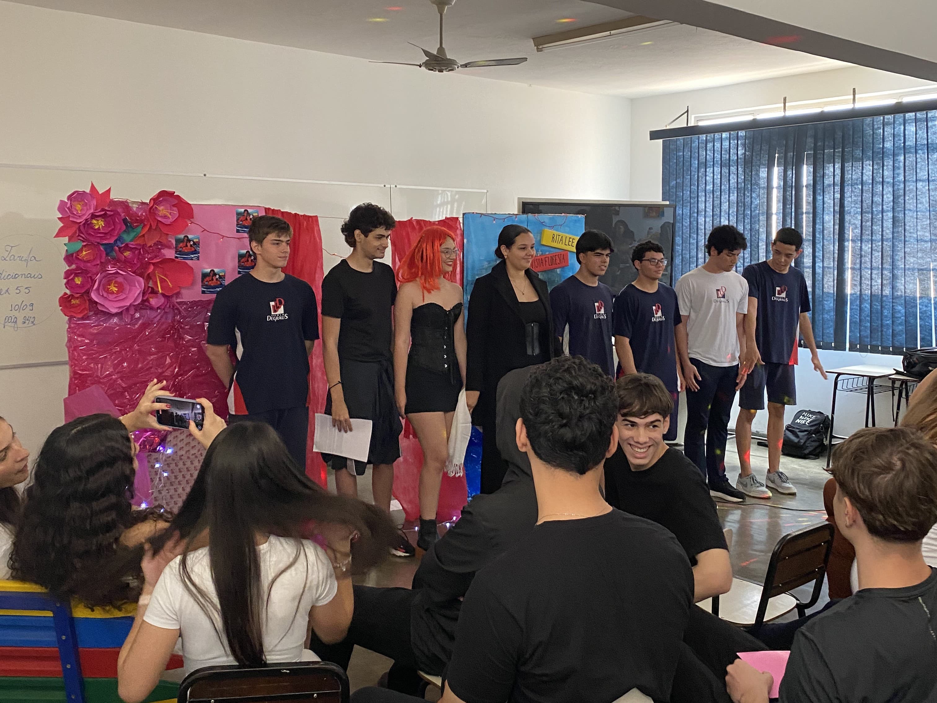Alunos da Escola Degraus apresentam uma encenação em sala de aula durante um evento escolar, enquanto colegas acompanham e registram a apresentação.