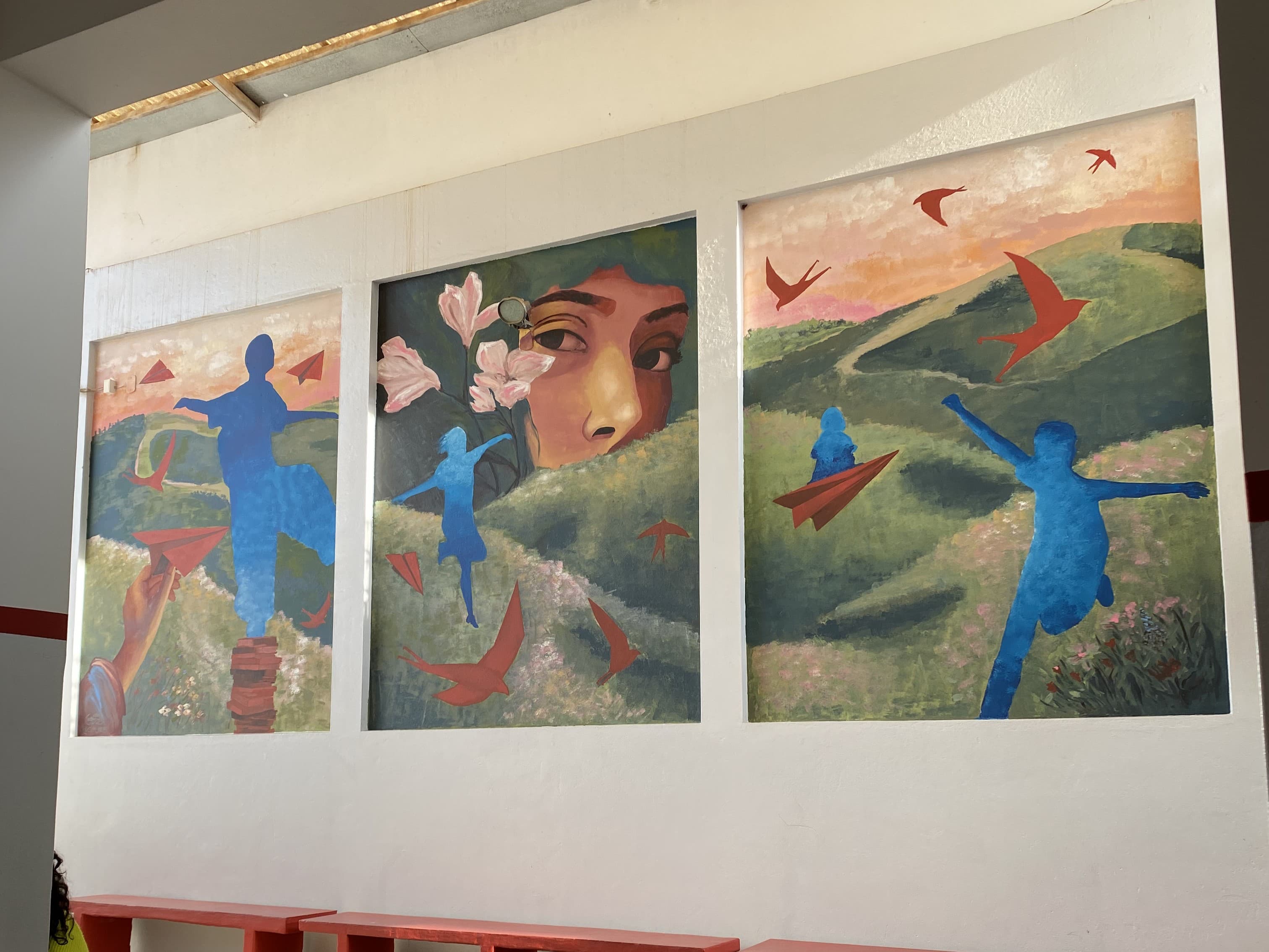 Painel artístico com três murais coloridos na Escola Degraus, retratando paisagens, figuras humanas em azul, pássaros vermelhos e um rosto ao centro.