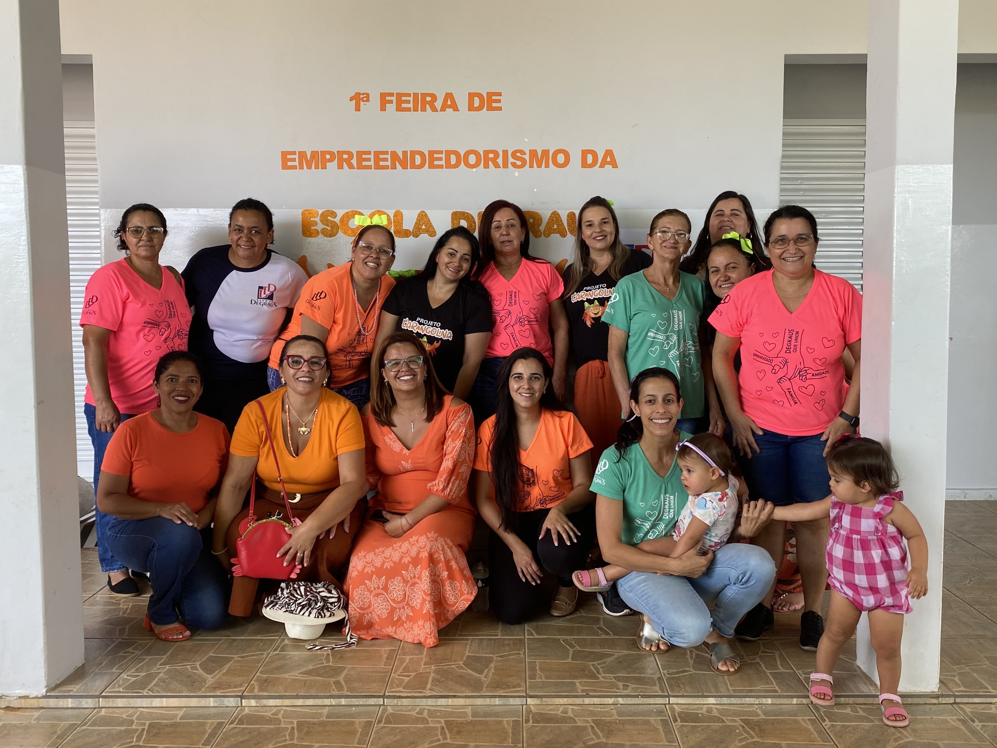 Foto de grupo celebrando a primeira feira de empreendedorismo de uma escola de educação infantil, com mulheres adultas e duas crianças sorrindo em frente a um banner do evento.