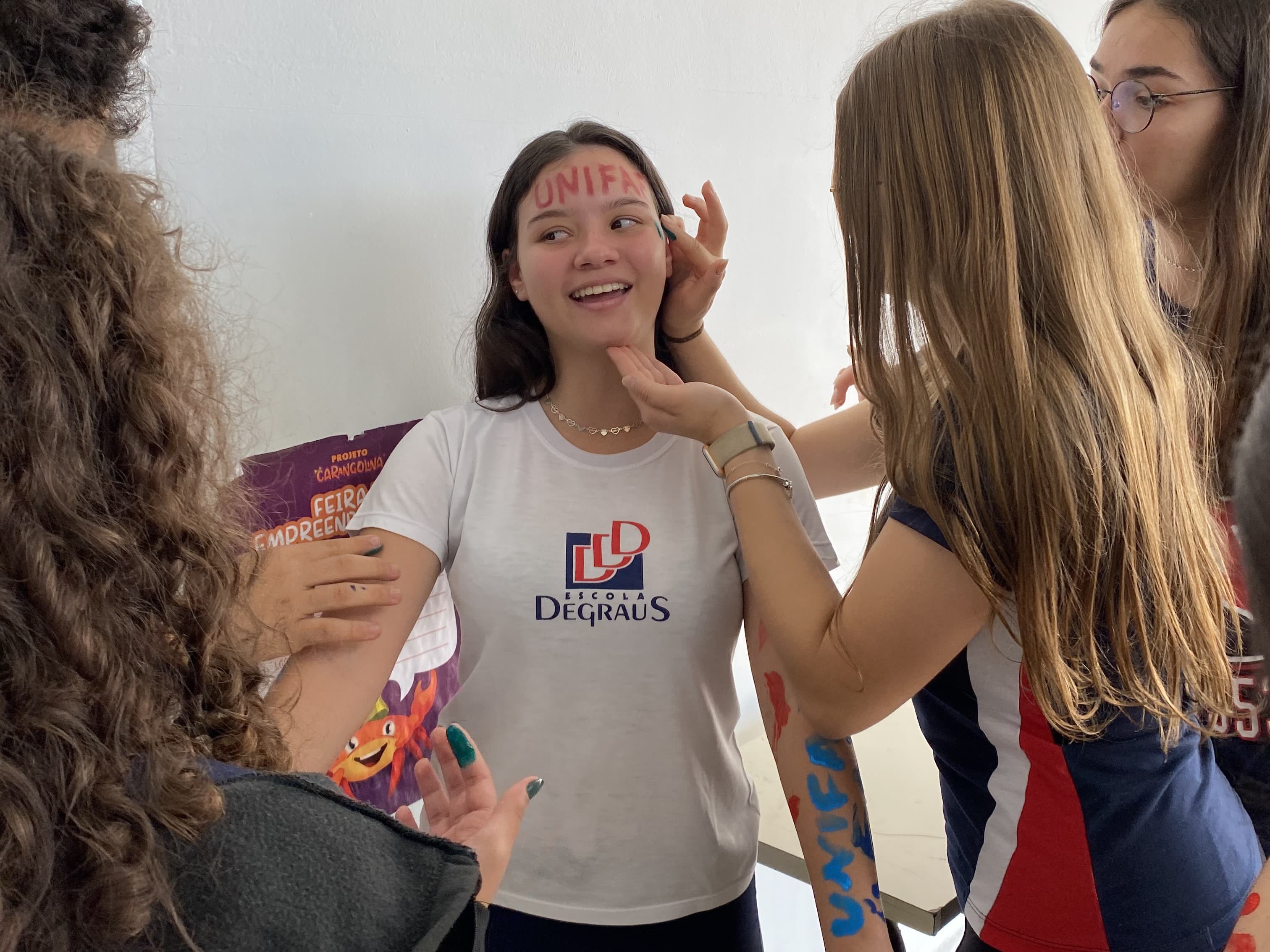 Aluna da Escola Degraus comemora sua aprovação no vestibular enquanto colegas escrevem mensagens de parabéns em seu rosto e braços.