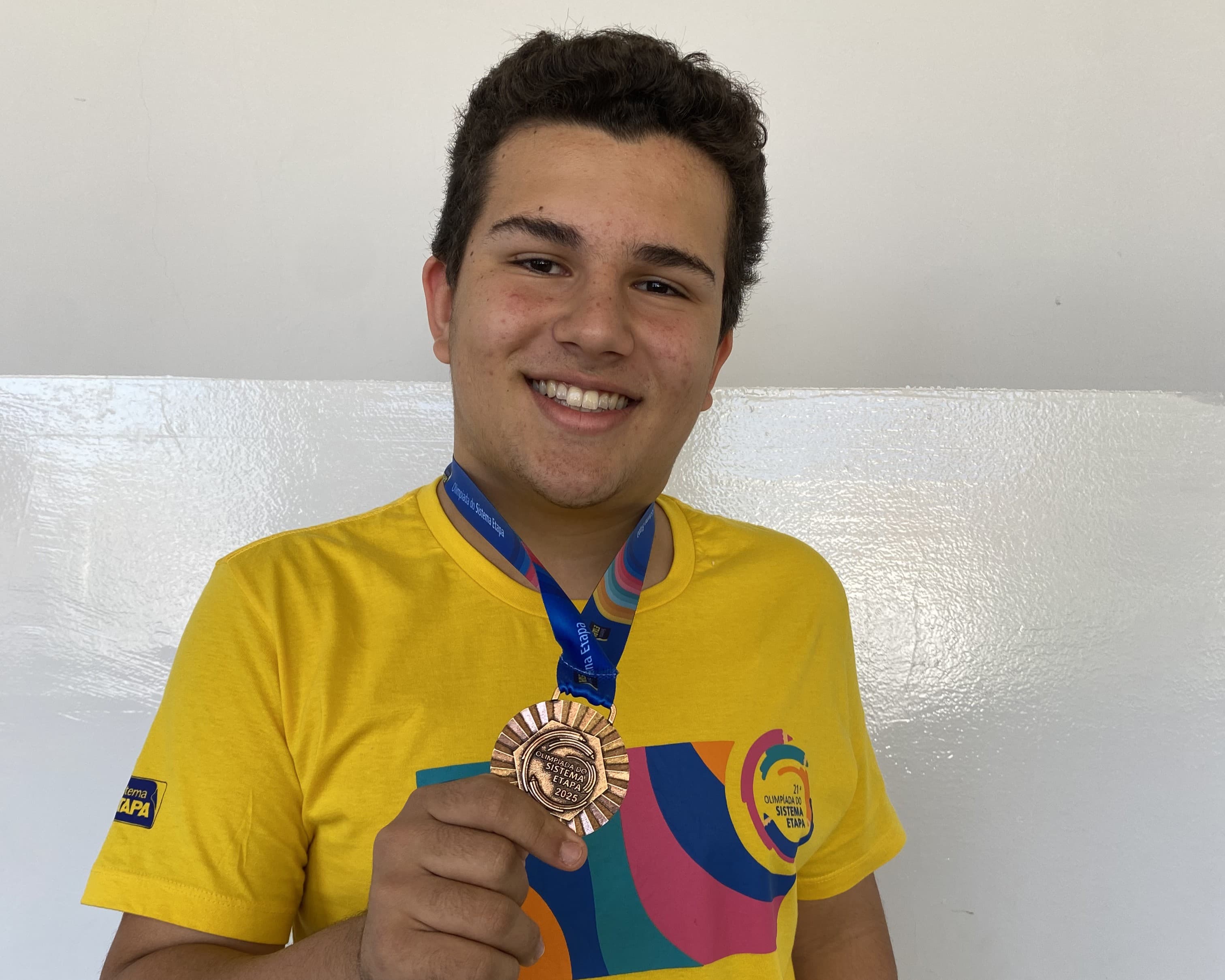 Estudante segura o troféu da 21ª Olimpíada do Sistema Etapa 2025, vestindo a camiseta amarela do evento.